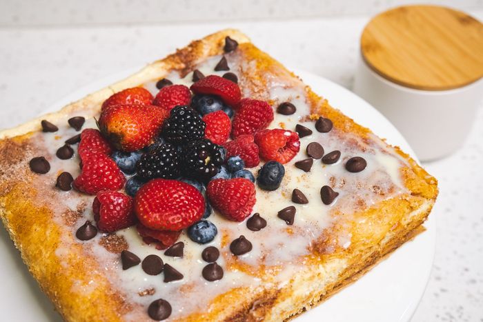 Frosted square pastry topped with mixed berries and chocolate chips beside a small cup of sauce