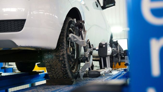 In un'officina, un'auto bianca è posizionata su un ponte sollevatore blu, con una macchina per l'allineamento delle ruote collegata alla ruota anteriore.