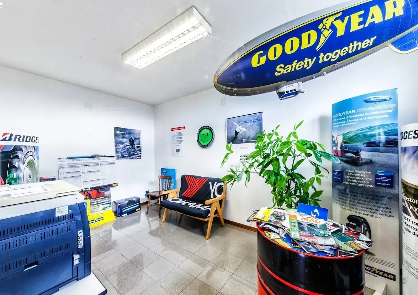 Area d'attesa del negozio di pneumatici Goodyear con poster, brochure, sedie, pianta in vaso e grande striscione del marchio.