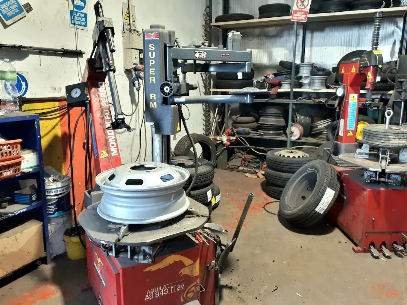 Interno di un'officina di pneumatici con una macchina per il montaggio pneumatici, ruote impilate e attrezzi su scaffali e banchi da lavoro.