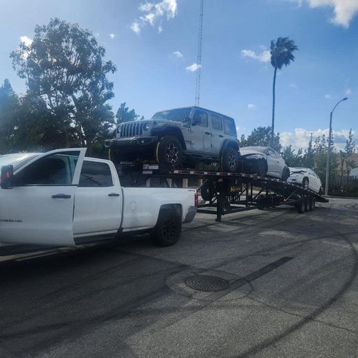 White truck hauling a trailer with a grey Jeep and two other cars on it, outdoors.