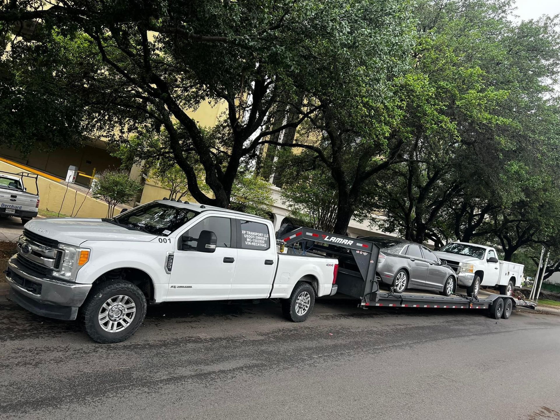 White pickup truck towing a trailer with two cars parked on a tree-lined street.