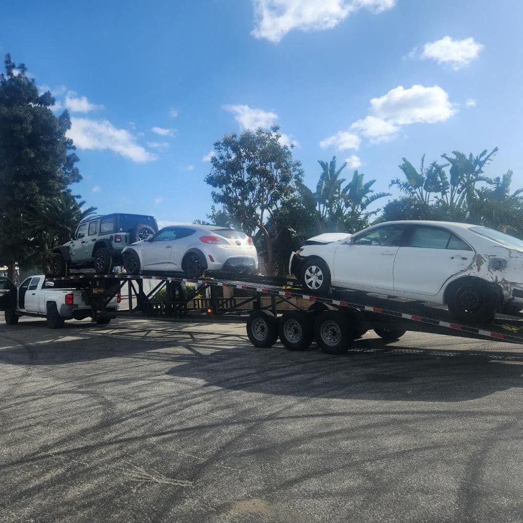 Three damaged cars on a trailer, loaded by a truck, under a blue sky.