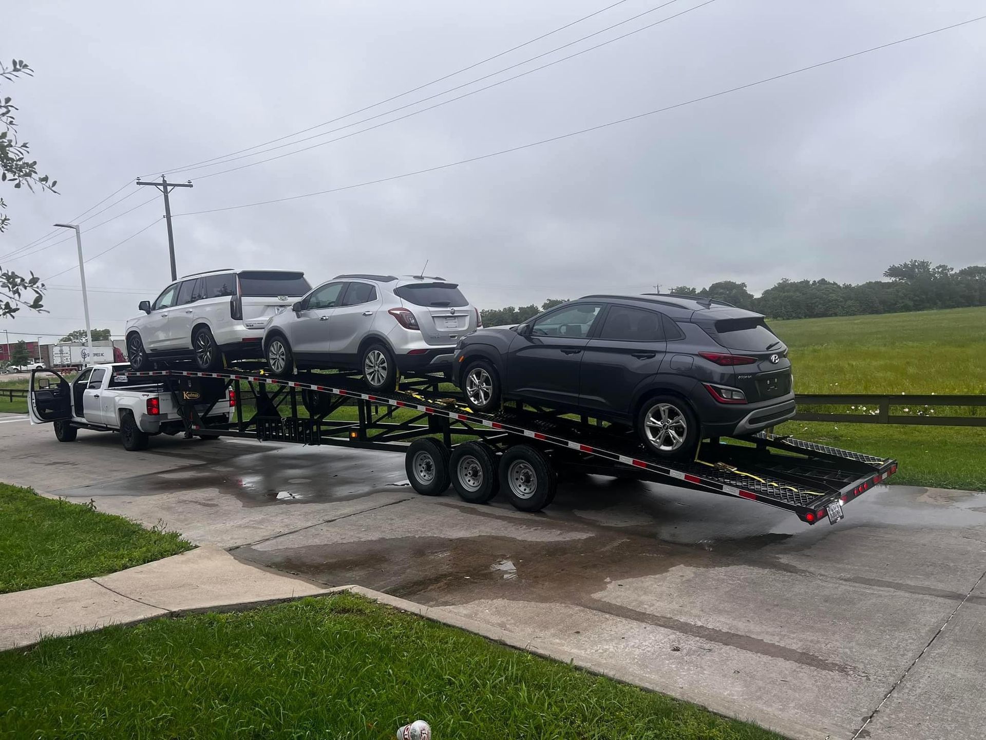 Car hauler truck carrying three SUVs on a wet road, overcast sky.