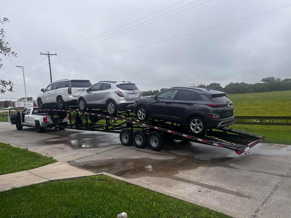 Car hauler truck carrying three SUVs on a wet road, overcast sky.