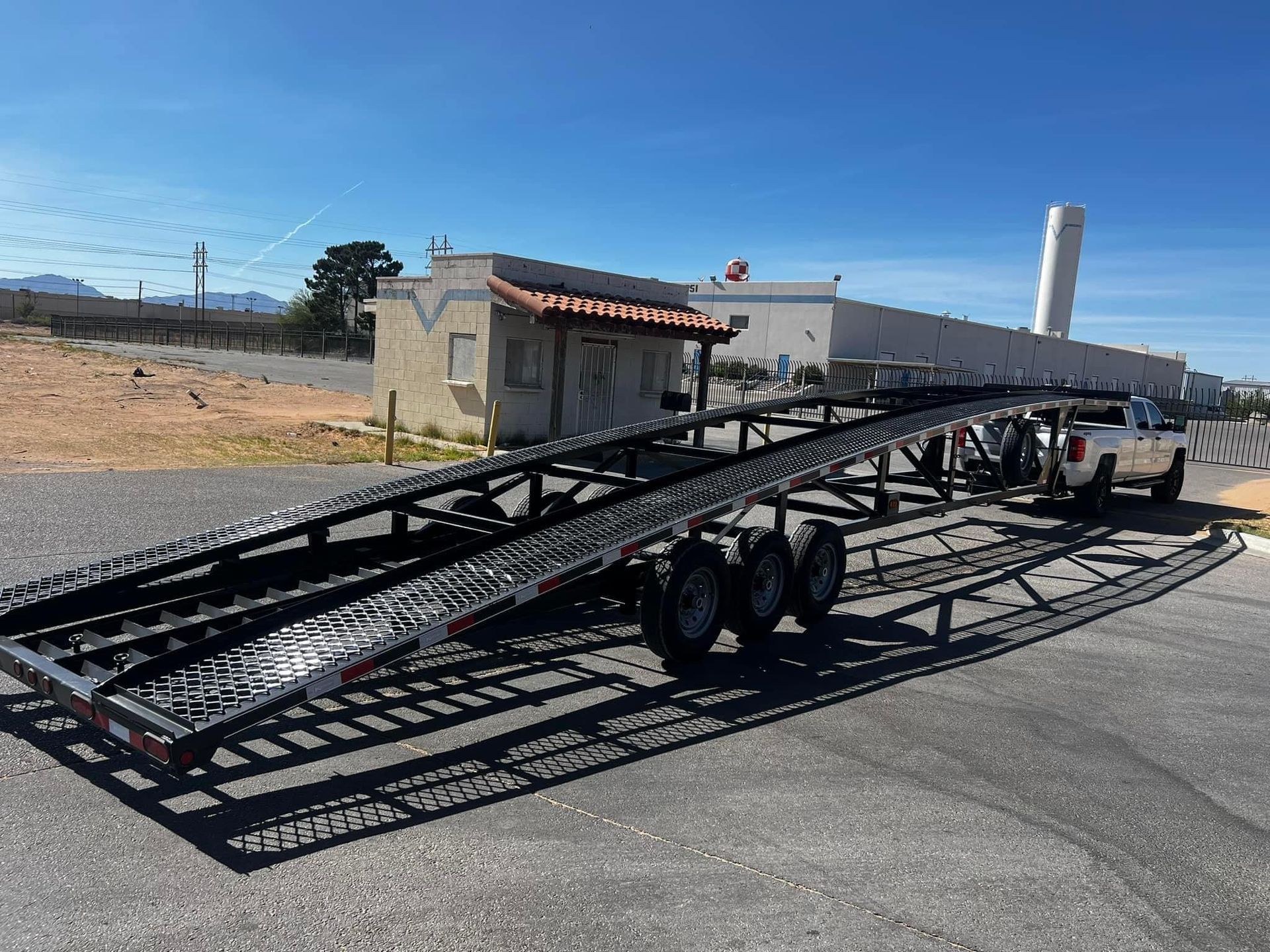 Black car hauler trailer hooked to a white pickup truck, parked outside a building on a sunny day.