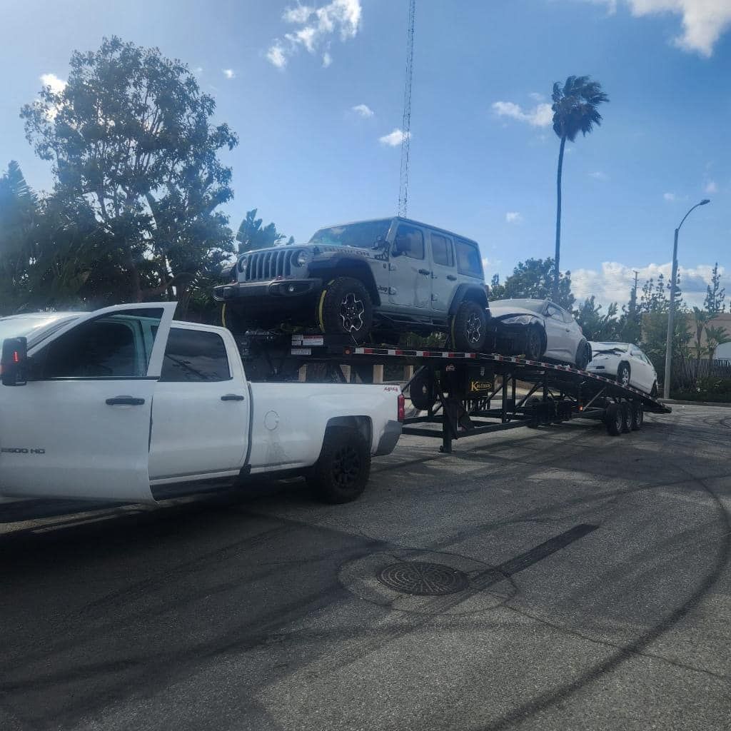 White truck towing a trailer with a Jeep and two cars on it, outdoors.