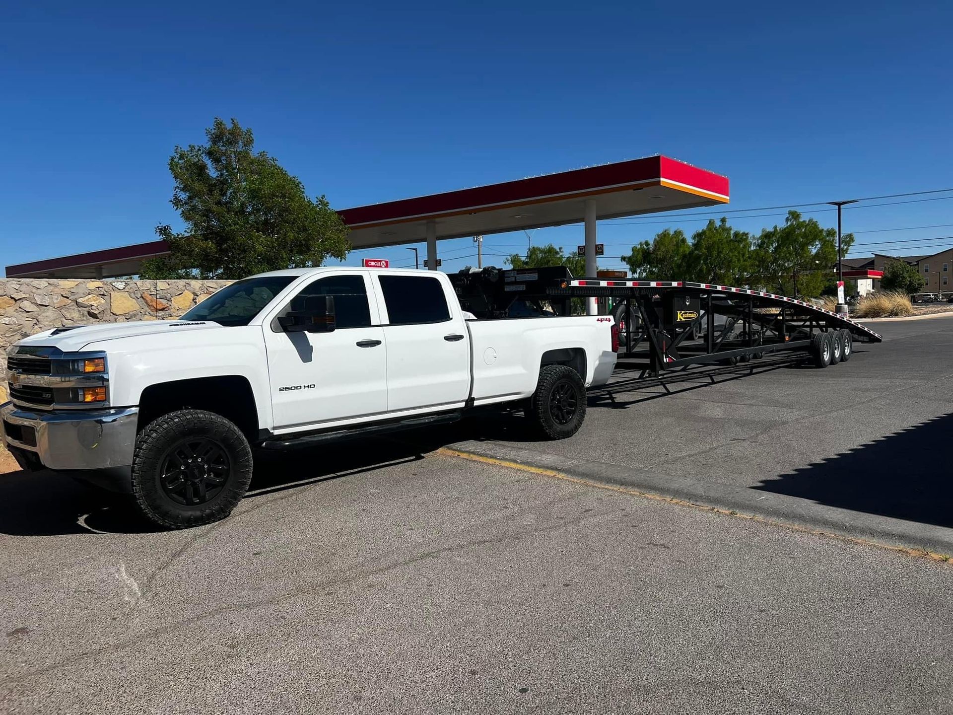 White pickup truck towing a car hauler at a gas station under a clear blue sky.