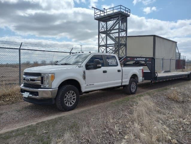 White pickup truck towing equipment on a flatbed trailer next to a tall metal structure.