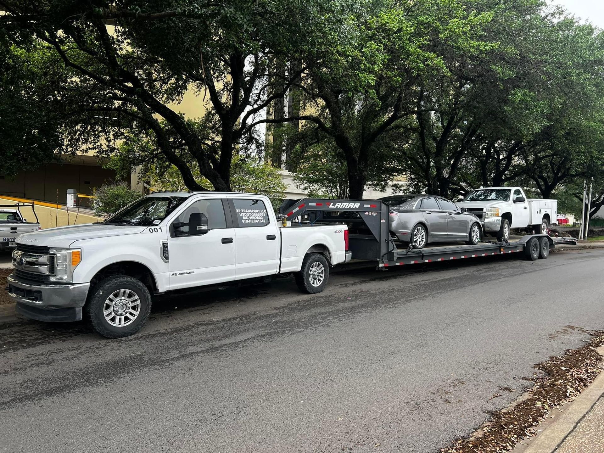 White pickup truck towing a trailer with two silver cars on a city street.