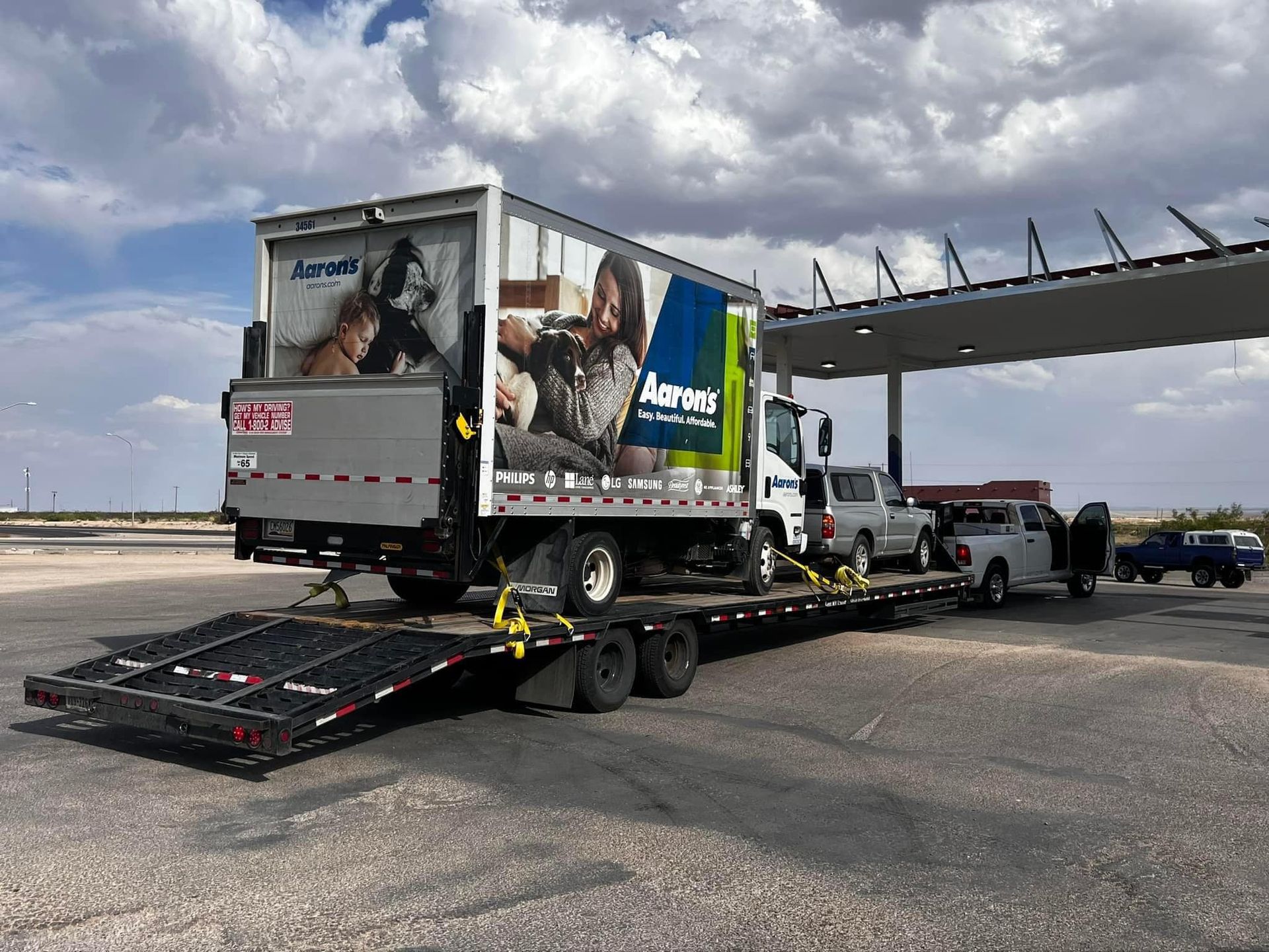 Box truck with Aspen banner on a flatbed trailer, parked at a gas station.