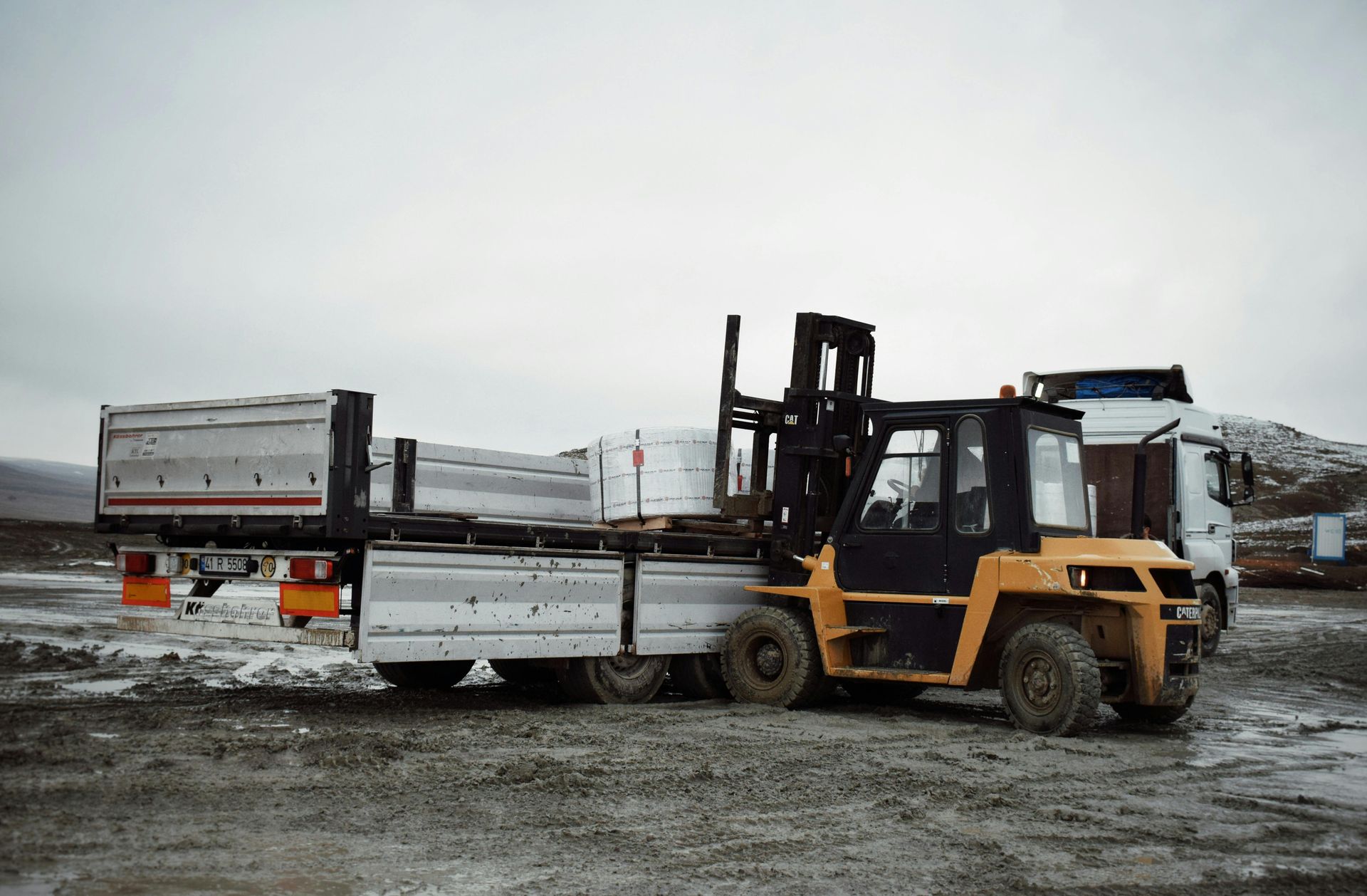 Yellow forklift loading cargo onto a flatbed trailer on a cloudy day.