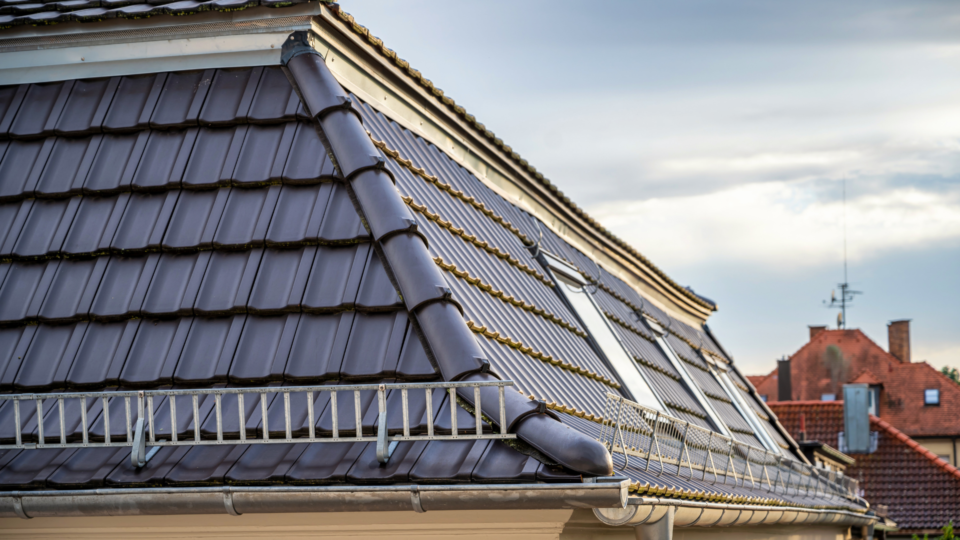 Dark gray tiled roof with skylights under a cloudy sky, with a distant brick building behind it