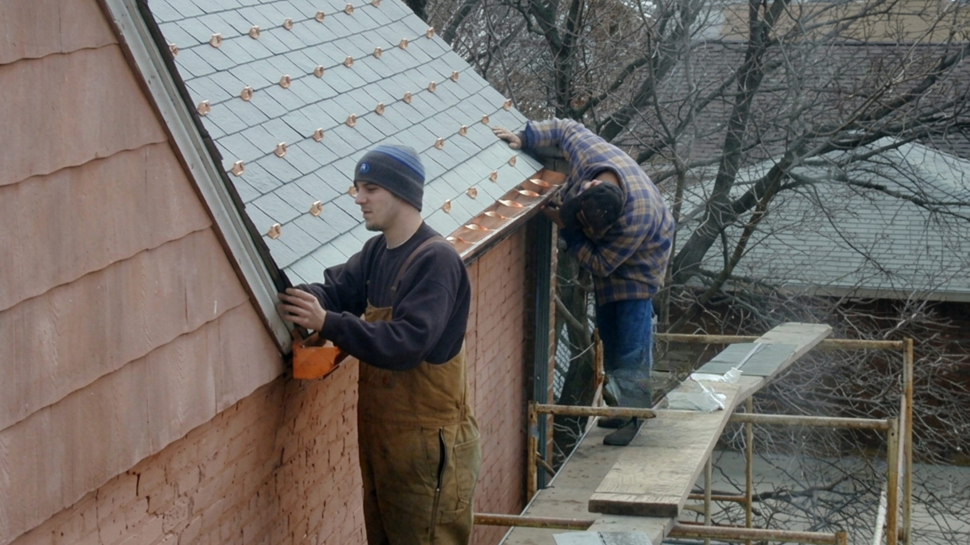 Two workers install shingles on a steep roof beside a wooden building.