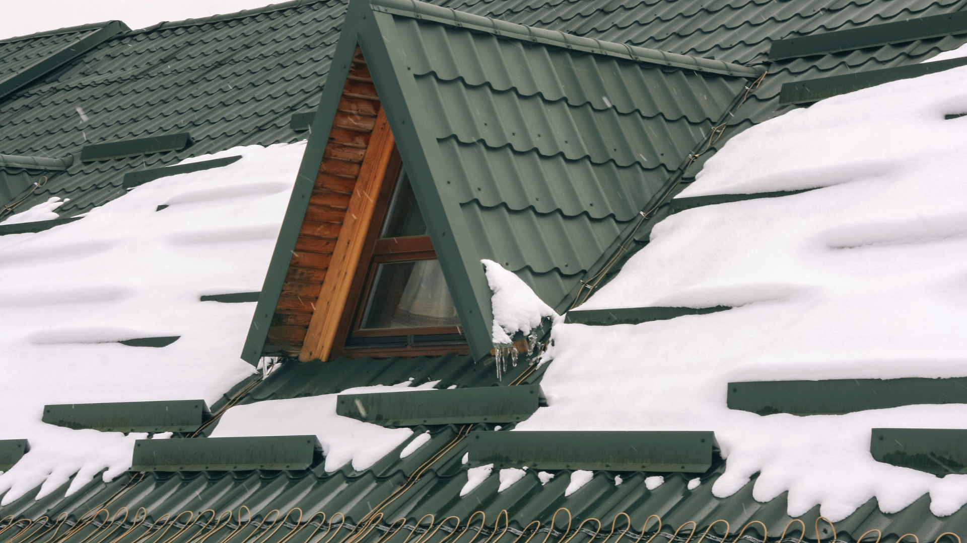 Snow-covered green metal roof with a small dormer window and wooden trim