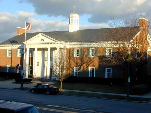 Brick civic building with white columns and flags, seen from across the street on a sunny day