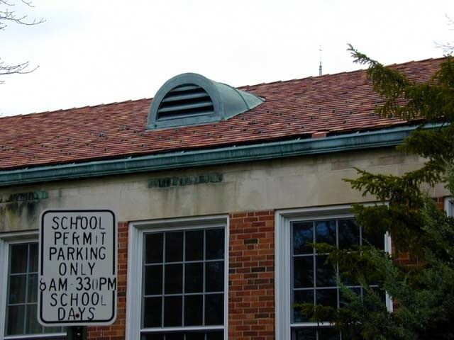School parking sign beside a brick building with red shingled roof and dormer window.