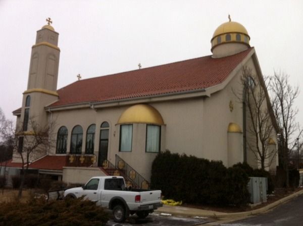 Small church with a red roof and gold domes, seen from the street with a white pickup truck parked outside.