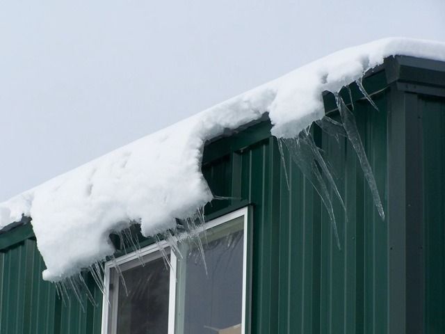 Snow piled on the roof of a green building with icicles hanging over a window