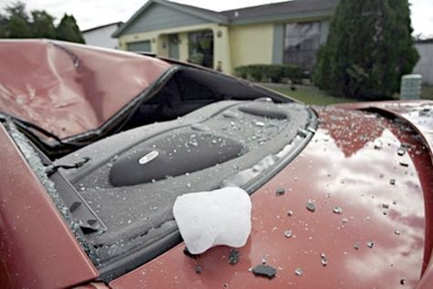 Red car with a smashed windshield and deployed airbag in front of a house.