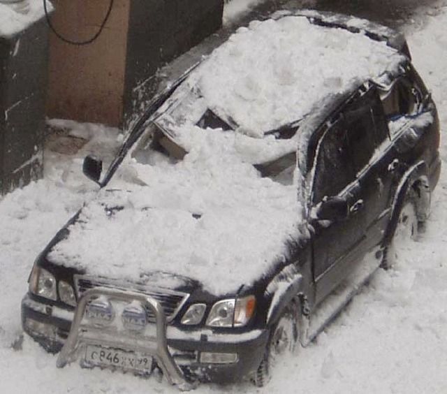 Black SUV covered in snow, parked beside a building in a snowy lot.