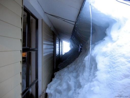 Snow piled high against a house, nearly reaching the roof along a narrow side walkway