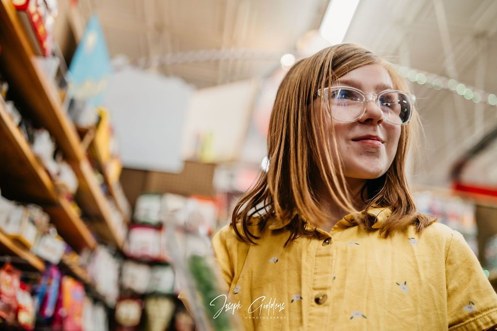 A person with reddish hair and glasses wearing a yellow shirt, looking off-camera while standing in a store aisle.