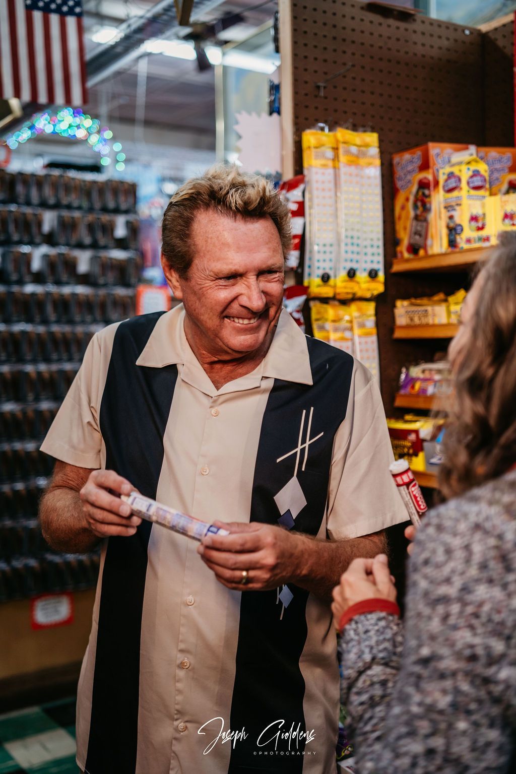 A smiling person wearing a bowling-style shirt holds a small product while interacting with someone in a retail store.