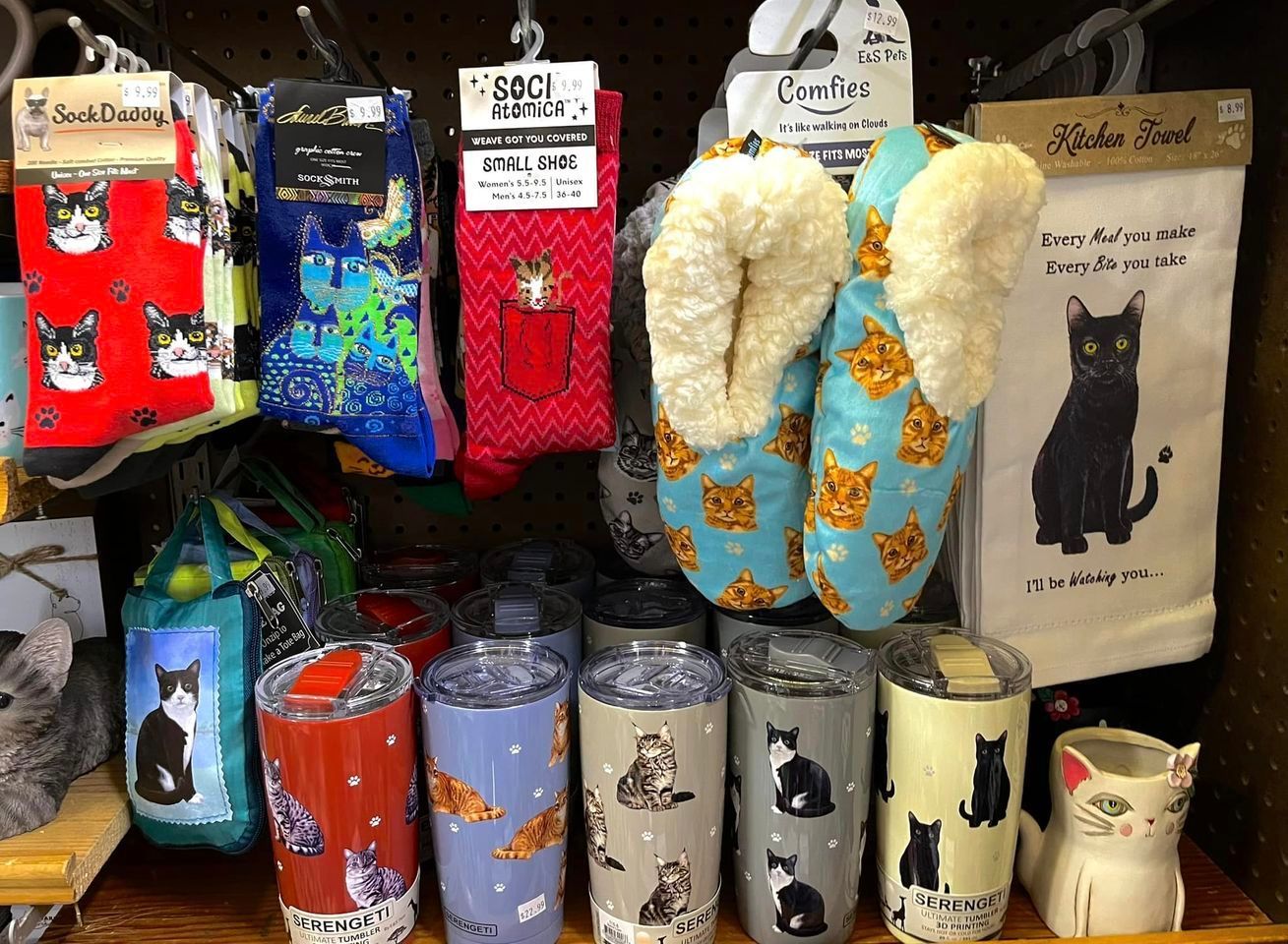 A display of cat-themed merchandise, including socks, fuzzy slippers, travel tumblers, and a tea towel with a black cat.