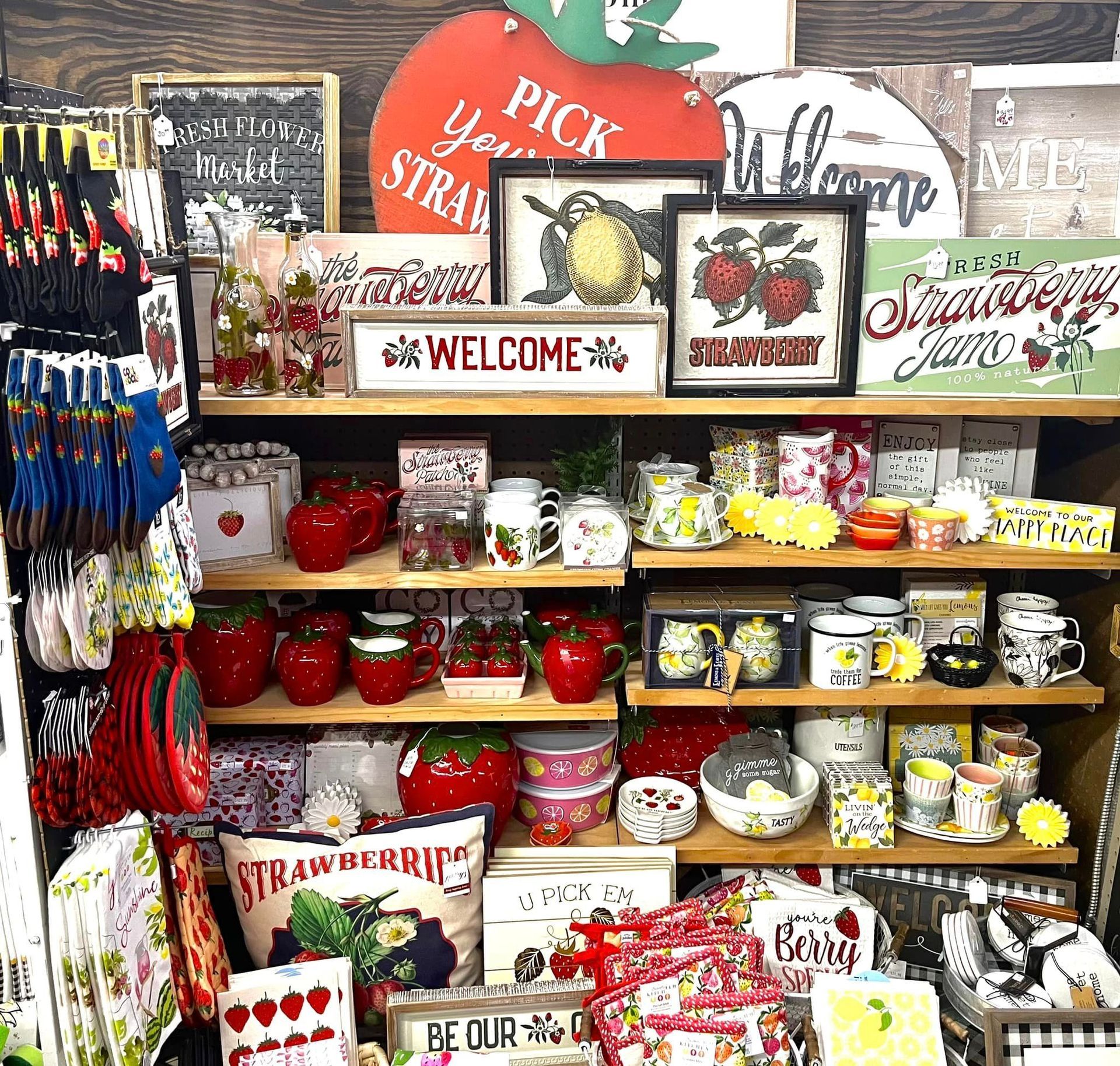 A retail display shelf filled with strawberry-themed home decor, including mugs, signs, kitchen towels, and small gifts.