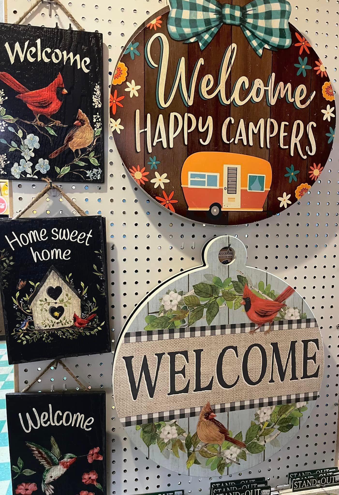 A pegboard display of five hanging wall signs featuring various floral, bird, and camper motifs with
