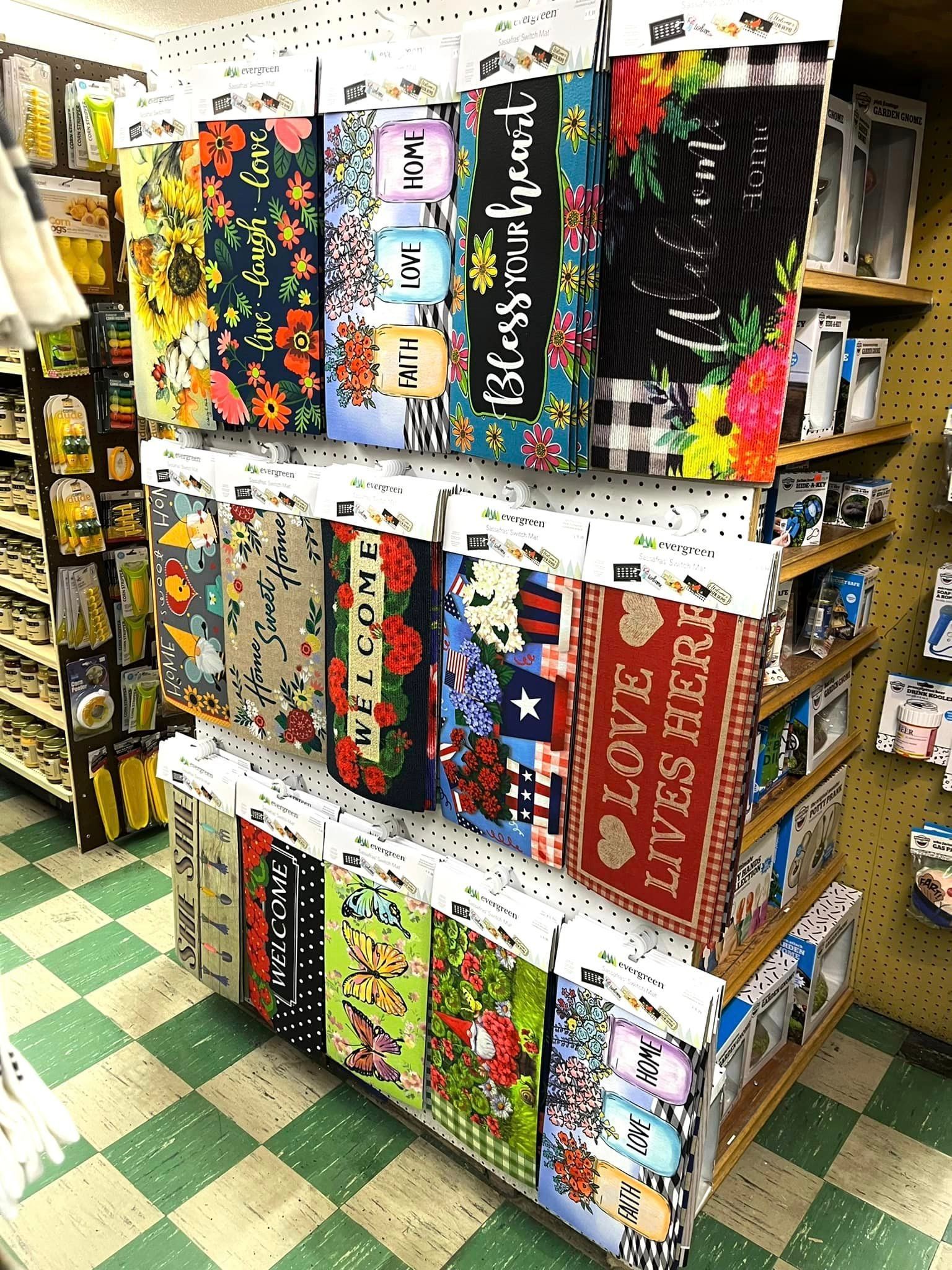 Shelves in a store display a variety of decorative welcome mats and signs with floral, seasonal, and patriotic themes.
