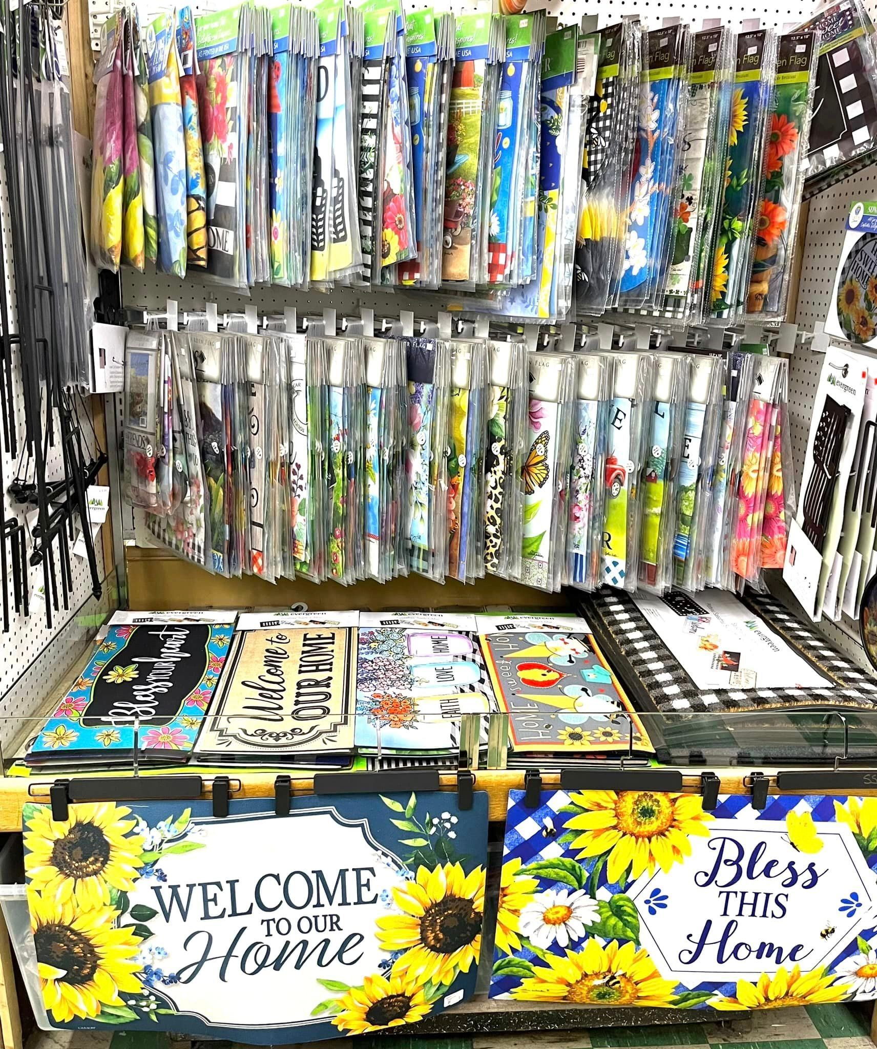 A display of colorful, decorative floor mats and vertical hanging signs featuring sunflowers and welcome messages.