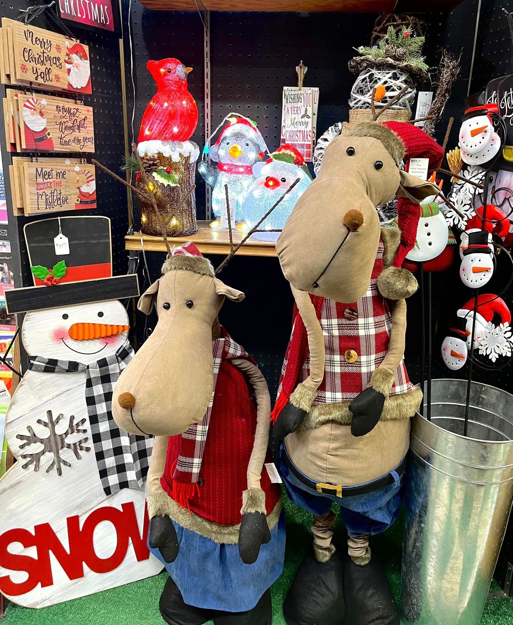 Two stuffed reindeer figurines stand on a store shelf next to a snowman sign and other holiday decor.