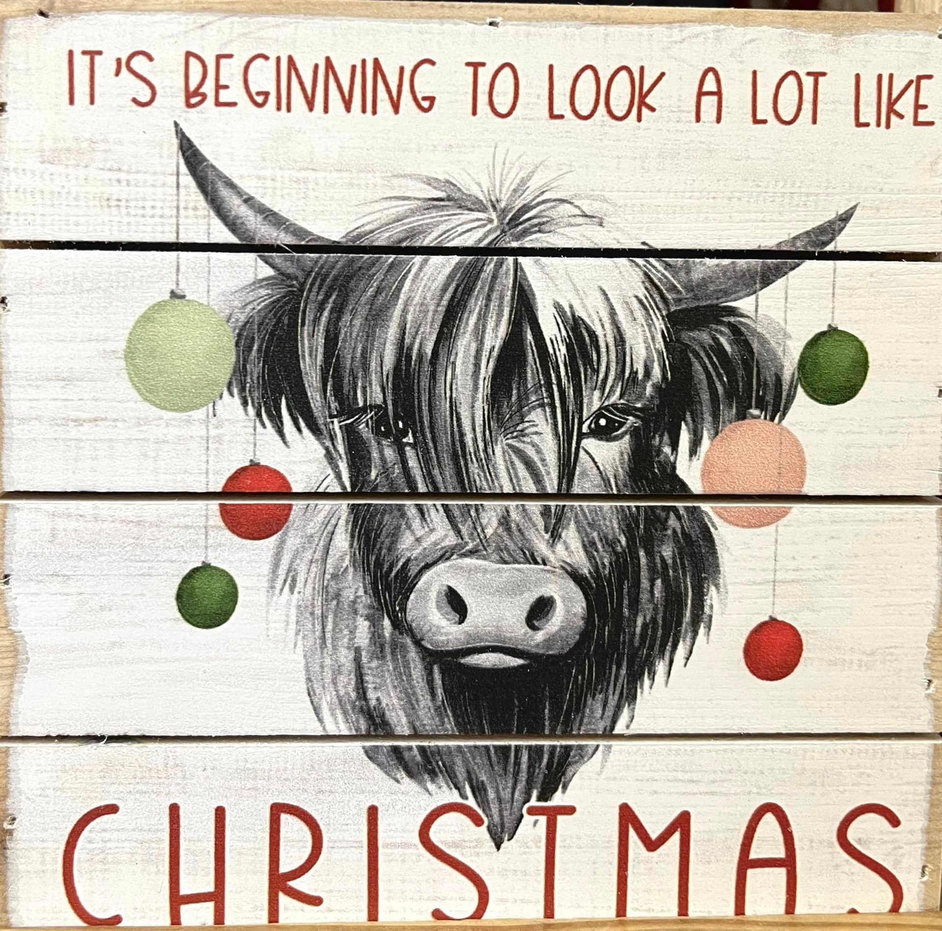 A decorative sign with a drawing of a Highland cow decorated with hanging colorful Christmas ornaments.