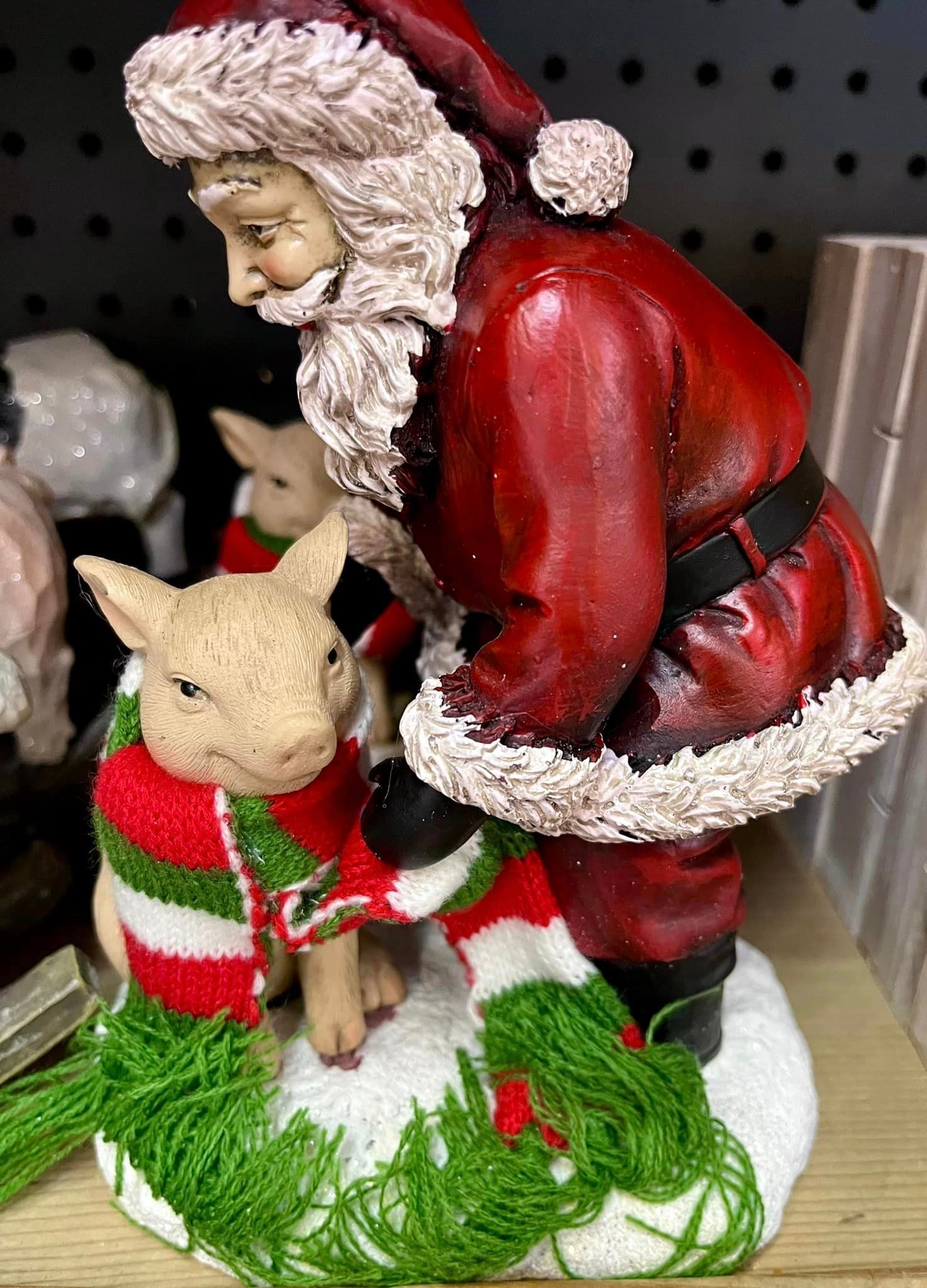 A Santa Claus figurine wearing a red suit leans over to wrap a red, white, and green striped scarf around a piglet.