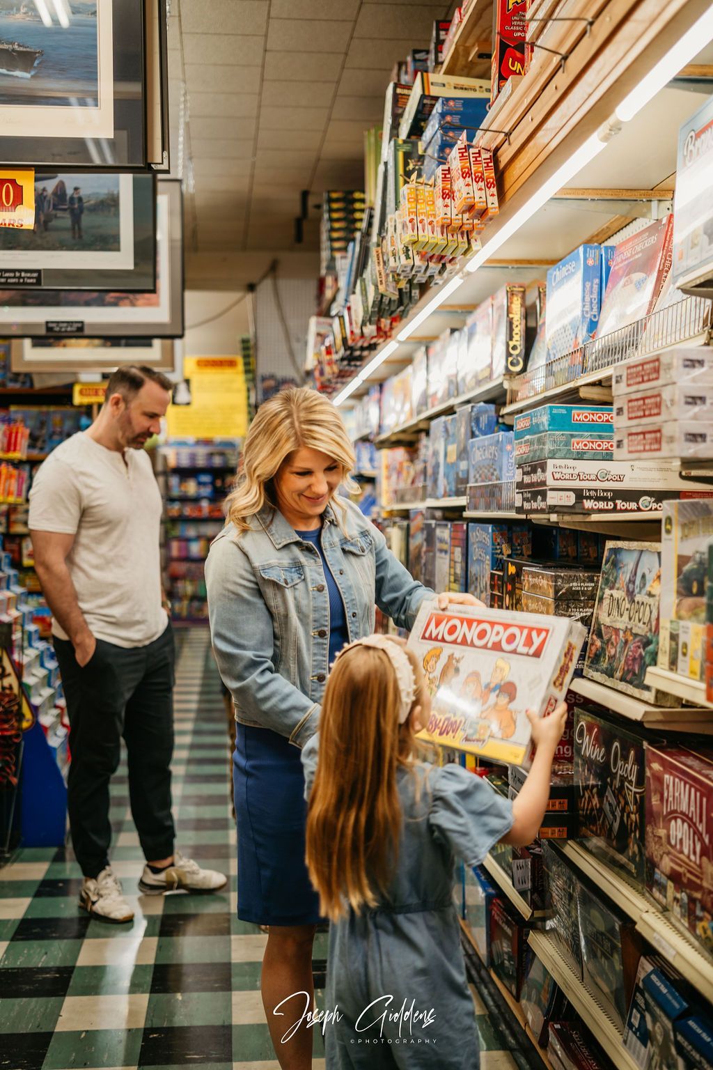 A family browses a toy store aisle as a child holds a Monopoly game box, with shelves of board games in the background.