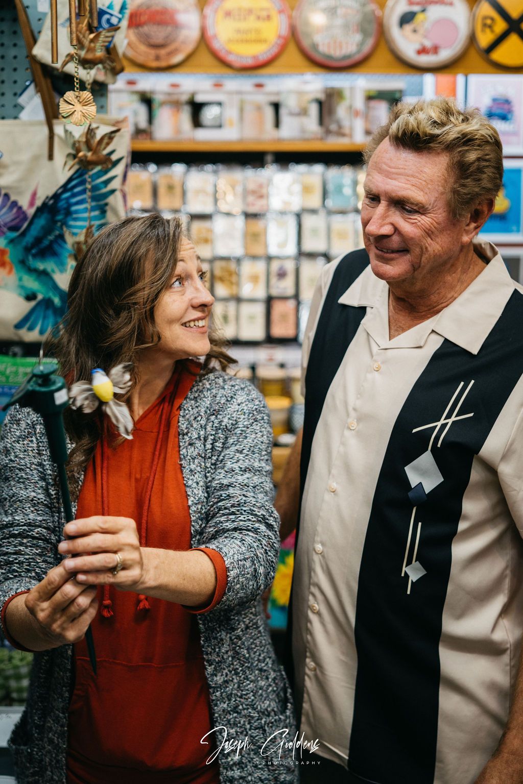 A person in a gray sweater and orange top examines a small bird ornament while a smiling person in a bowling shirt looks on.
