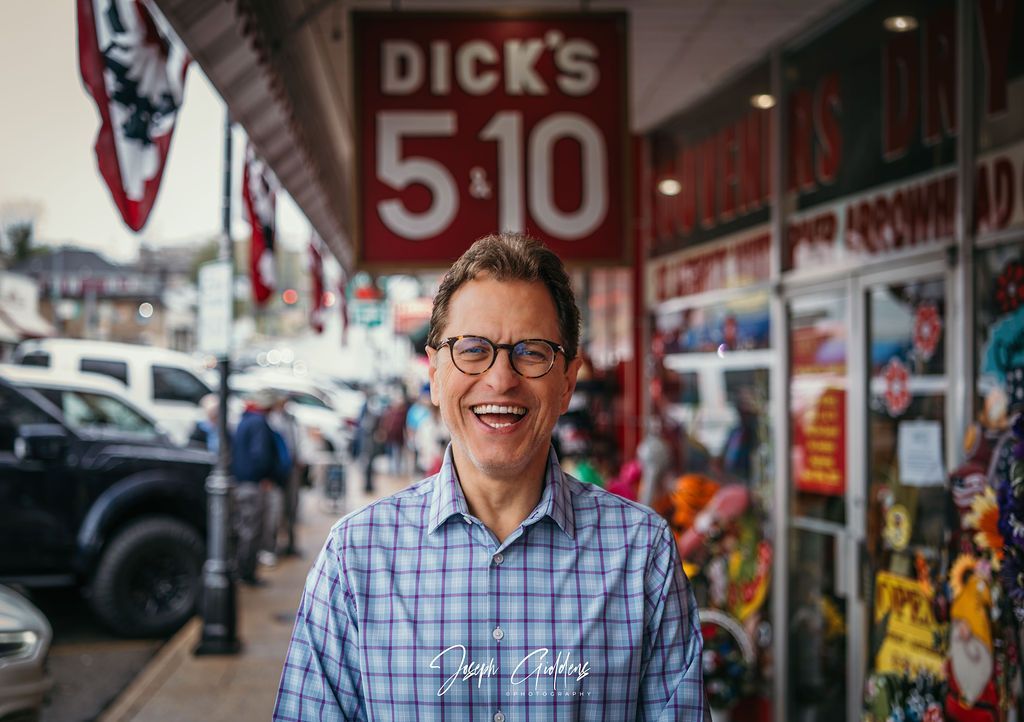 A smiling person in a blue plaid shirt stands outside 