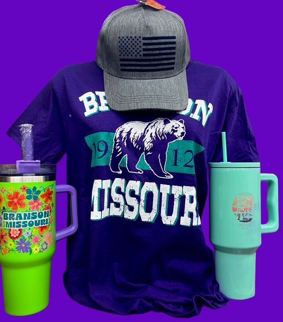 A grey hat with a flag patch, a purple Branson Missouri bear t-shirt, and two tumblers against a purple background.