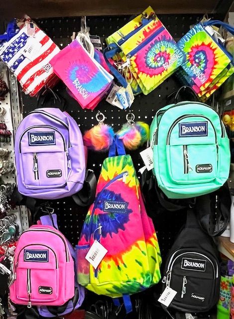 Display of colorful backpacks and small pouches for sale, featuring tie-dye, neon, and flag patterns on a store wall.