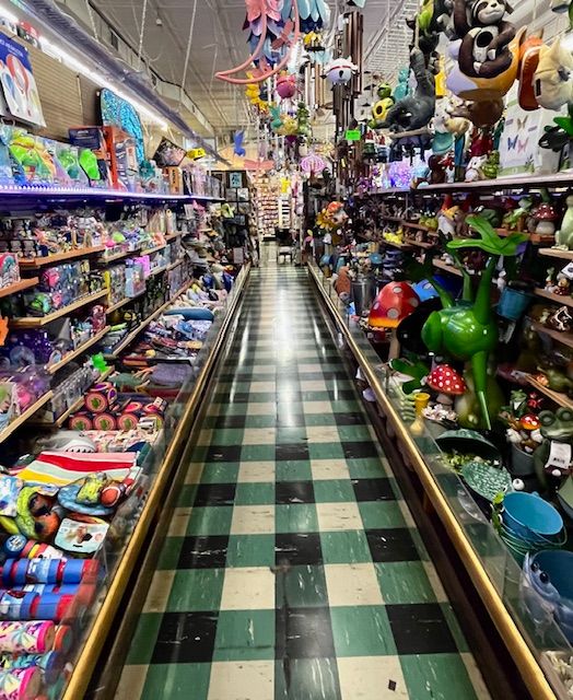 An aisle in a gift shop with green and black checkered flooring, featuring shelves filled with toys, gifts, and ornaments.