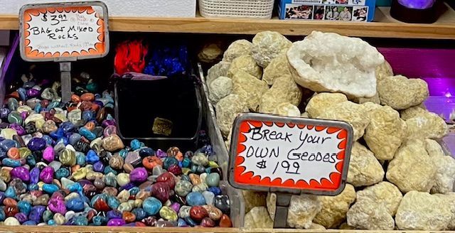 A retail display featuring a bin of assorted polished, colorful tumbled stones next to a bin of uncracked, natural geodes.