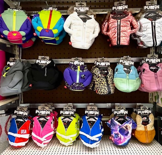 Three rows of colorful, insulated Puffin beverage jackets hang on pegs against a dark, perforated store display board.