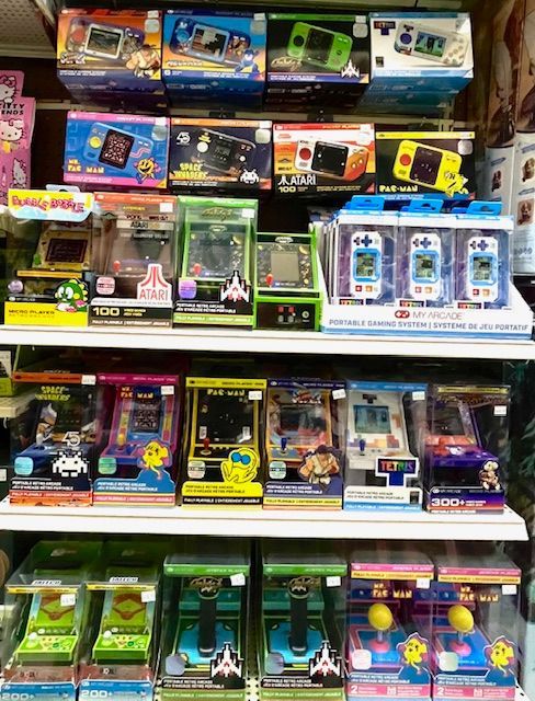 Store shelf stacked with various colorful, boxed retro-style handheld gaming consoles and mini arcade systems.