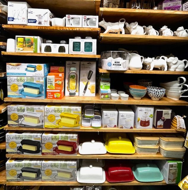 Wooden store shelves stocked with various kitchenwares, including butter dishes, white ceramic animal figures, and bowls.