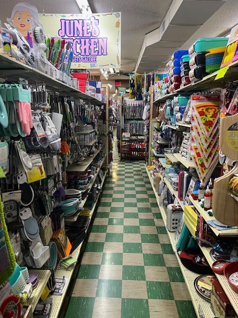 A narrow store aisle features shelves packed with kitchenware and home goods, with a checkered floor and a store sign.