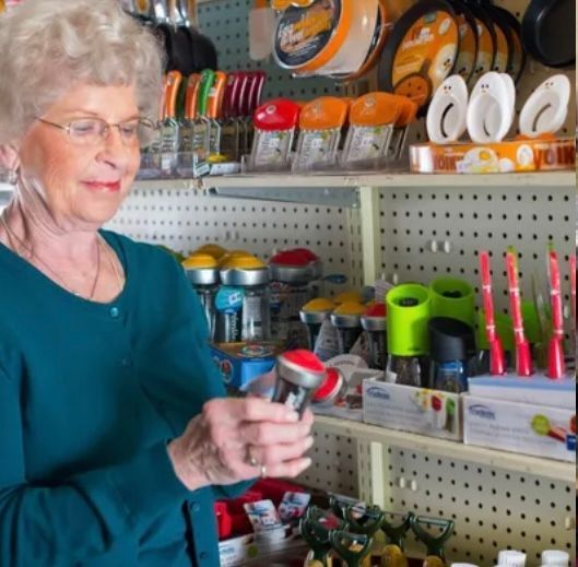 A person in a teal sweater examines a kitchen tool while standing in a store aisle filled with colorful cooking utensils.