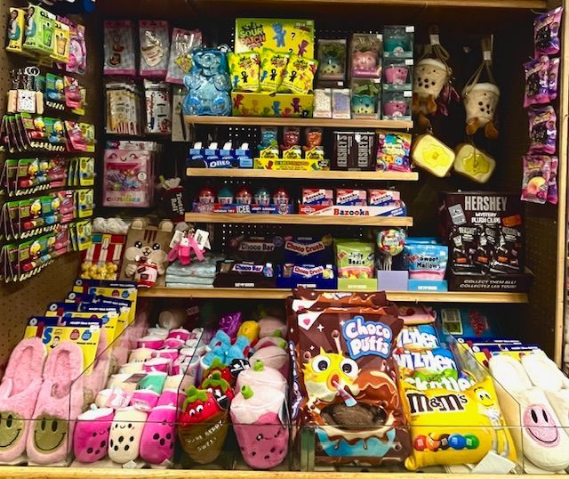 A retail shelf display filled with colorful packaged snacks, plush toys, and patterned house slippers.