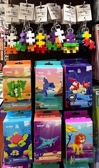 Display of Plus-Plus building toy sets in boxes and small keychain versions hanging on hooks in a retail store.